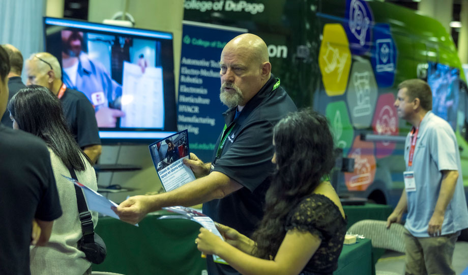 COD Welding Professor Tom Ellis hands a COD Manufacturing program flyer to a female who visited his booth at the International Manufacturing Technology Show