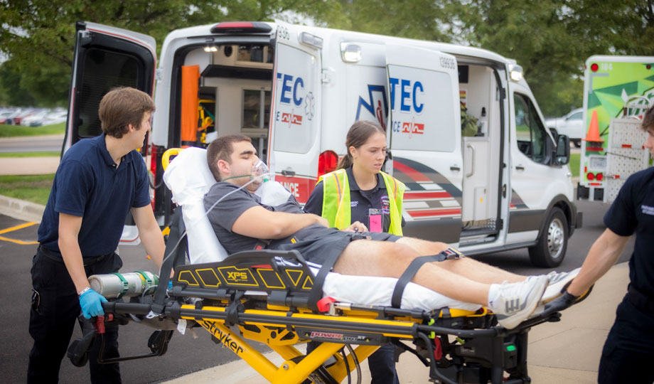 Two EMT students push a stretcher with a student strapped in.