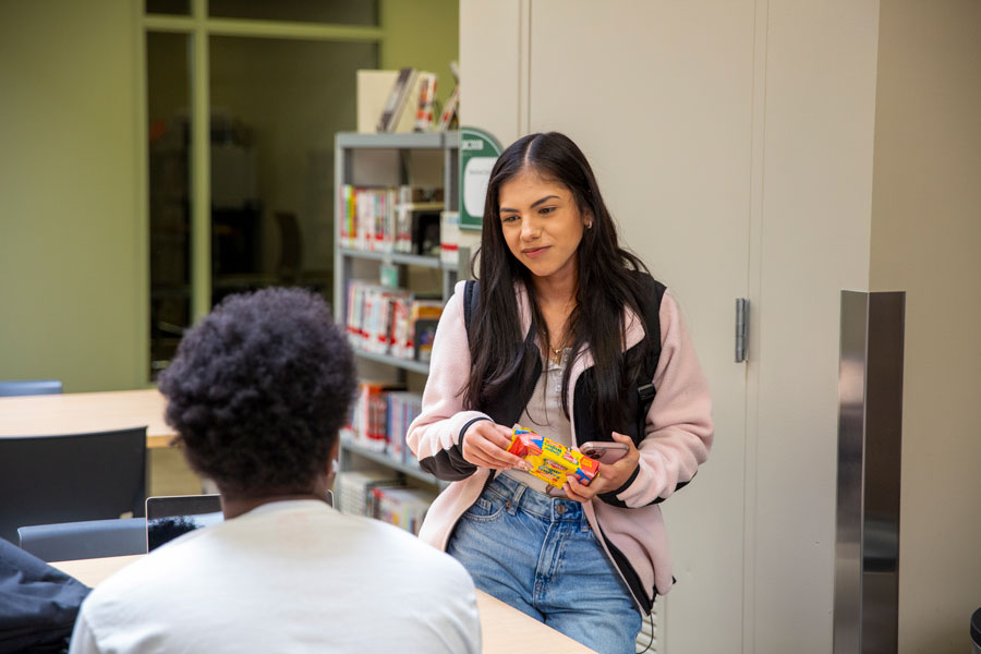 Student in pink jacket talking to sitting student.